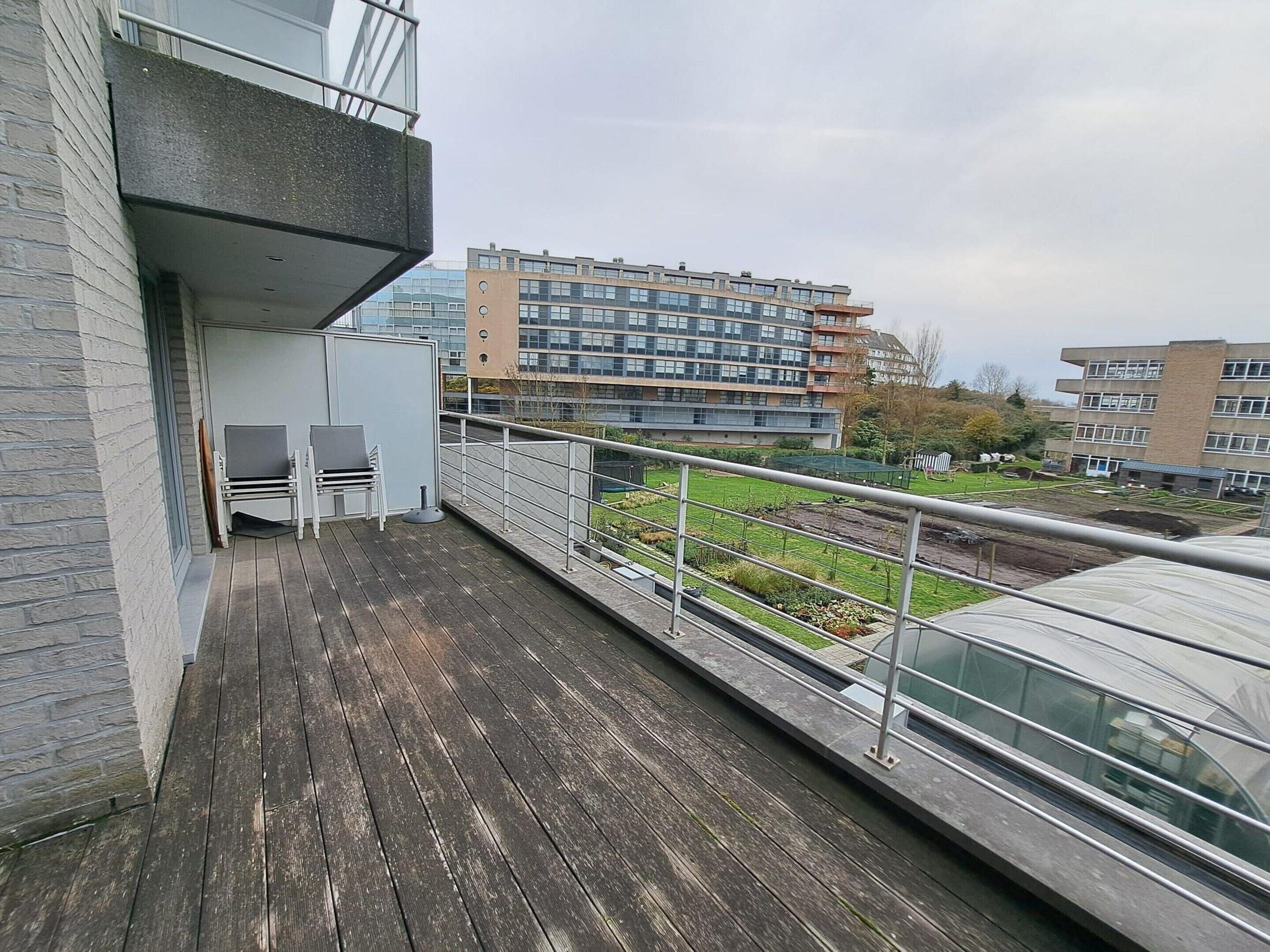 A balcony with a wooden floor, metal railing, and a view of a building with multiple windows and a garden area.