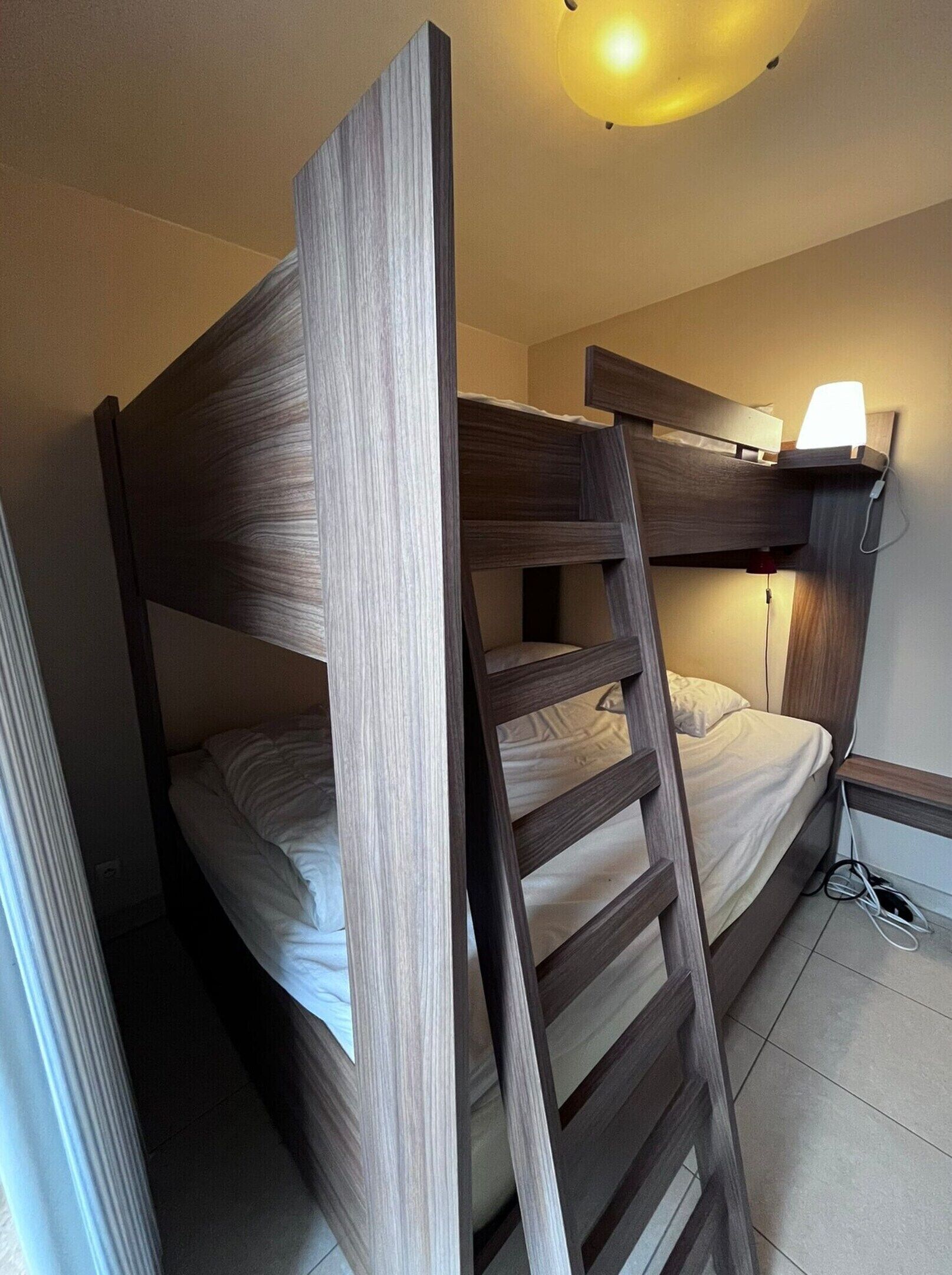A bunk bed room with a ladder, a bedside table with a lamp, and a power outlet.