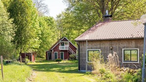 En træhytte med rødt tag, en balkon og et lille skur i et skovområde.