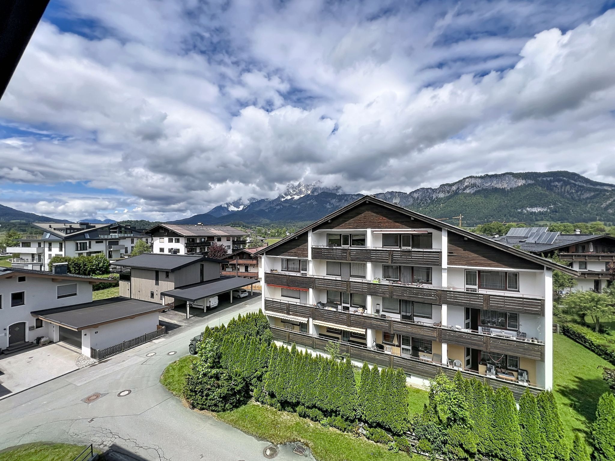 Un immeuble moderne à plusieurs étages, doté de balcons, entouré de verdure et avec des montagnes en arrière-plan.