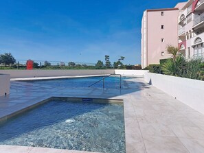 Ein Schwimmbecken mit Nichtschwimmerbereich, umgeben von einer Fliesenveranda und einem Gebäude mit Balkonen.