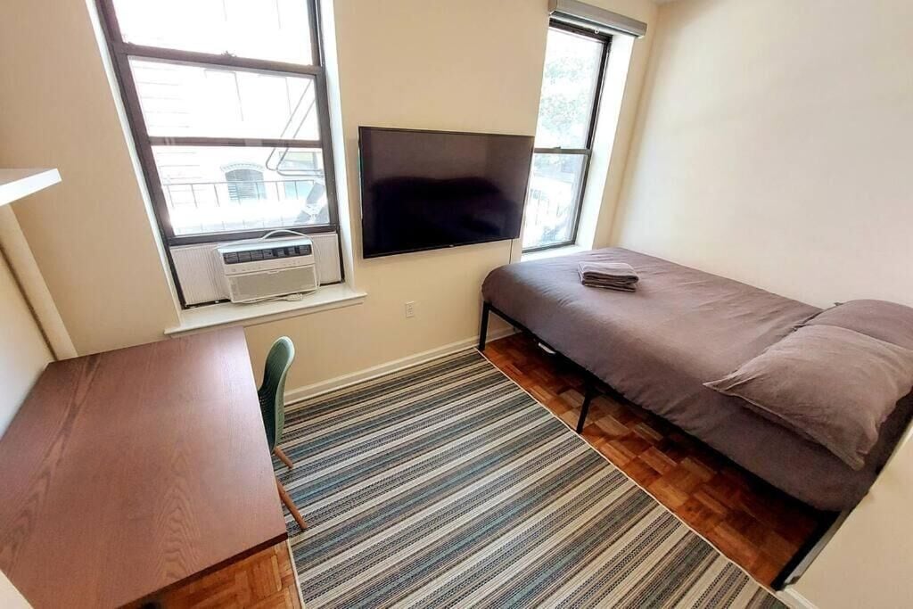 Ein kleines Zimmer mit einem Bett, einem Schreibtisch, einem Stuhl, einem Fernseher und einem Fenster.