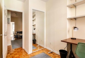 A room with a wooden floor, a desk with a lamp, a green chair, and a closet with shelves. - Large 2 Bedrooms with Queen Beds (NEW YORK)