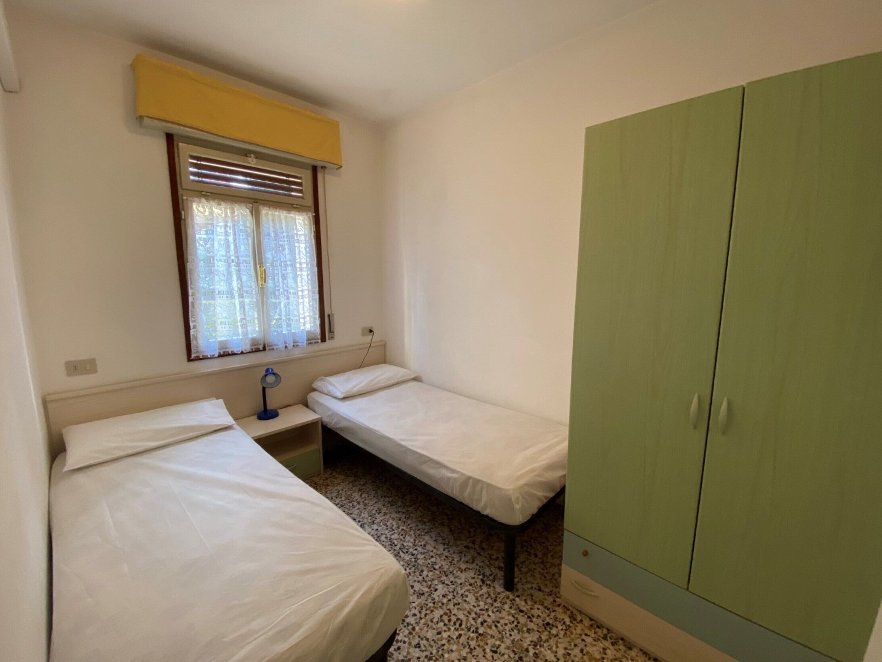 A small, single-room accommodation with two single beds, a bedside table with a lamp, a window with blinds, and a green wardrobe.