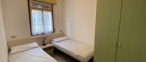 A small, single-room accommodation with two single beds, a bedside table with a lamp, a window with blinds, and a green wardrobe.