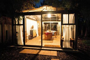 A glass-enclosed structure with a wooden interior, a table, chairs, and a hanging light fixture.