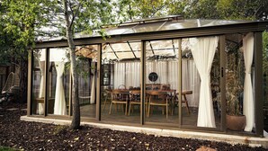 Een glazen constructie met een eettafel en stoelen, omringd door een tuin.