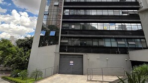 Un edificio moderno con fachada de vidrio reflectante, entrada porticada y patio empedrado con áreas verdes.