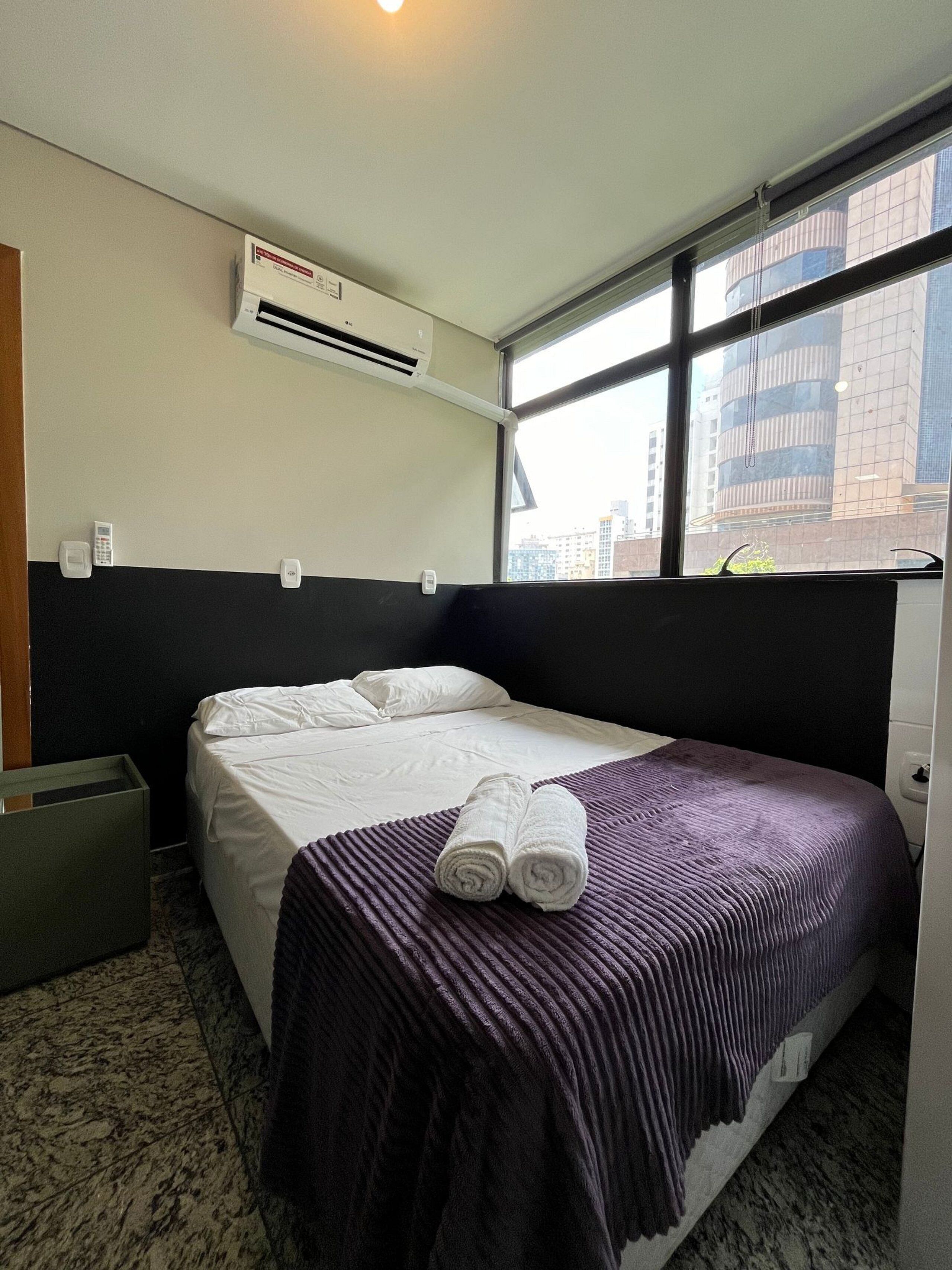 Habitación de hotel con cama, ventana con vista a la ciudad, equipo de aire acondicionado y teléfono junto a la cama.
