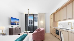 Een moderne hotelkamer met een bed, een bureau, een roze stoel, een keuken met houten kasten, een marmeren aanrechtblad en uitzicht over de stad.