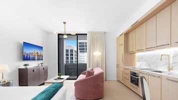 Une chambre d’hôtel moderne avec un lit, un bureau, une chaise rose, une cuisine dotée de meubles en bois, un plan de travail en marbre et une vue sur la ville.
