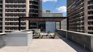 An outdoor terrace with a covered seating area, a view of a modern building, and a closed shutter door. - Sosuite | Queen Room, High Floor w Desk, Roof Deck (Philadelphia)