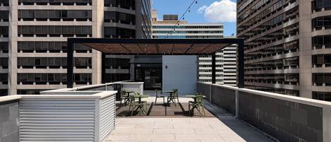 An outdoor terrace with a covered seating area, a view of a modern building, and a closed shutter door.
