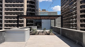 An outdoor terrace with a covered seating area, a view of a modern building, and a closed shutter door.