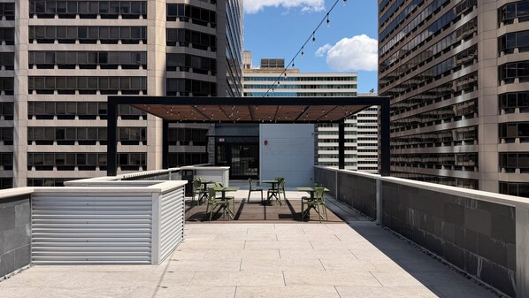 An outdoor terrace with a covered seating area, a view of a modern building, and a closed shutter door.