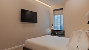 A modern hotel room with a bed, a desk, a chair, a flat-screen TV, and a window with a city view. - Sosuite | Queen Room, High Floor w Desk, Roof Deck (Philadelphia)