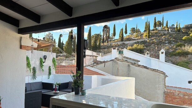 A balcony with a view of a hilly landscape, a seating area with cushions, and a small table with a plant.