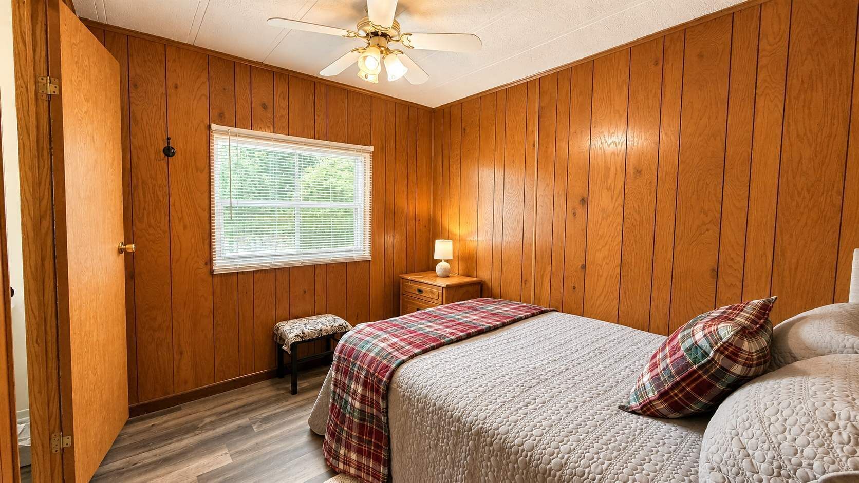 Un dormitorio con paneles de madera, una cama con colcha a cuadros, una mesita de noche con lámpara y una ventana con persianas.