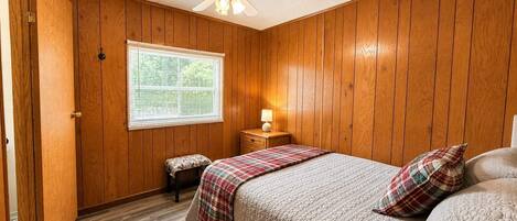 A bedroom with wooden paneling, a bed with a plaid bedspread, a nightstand with a lamp, and a window with blinds.
