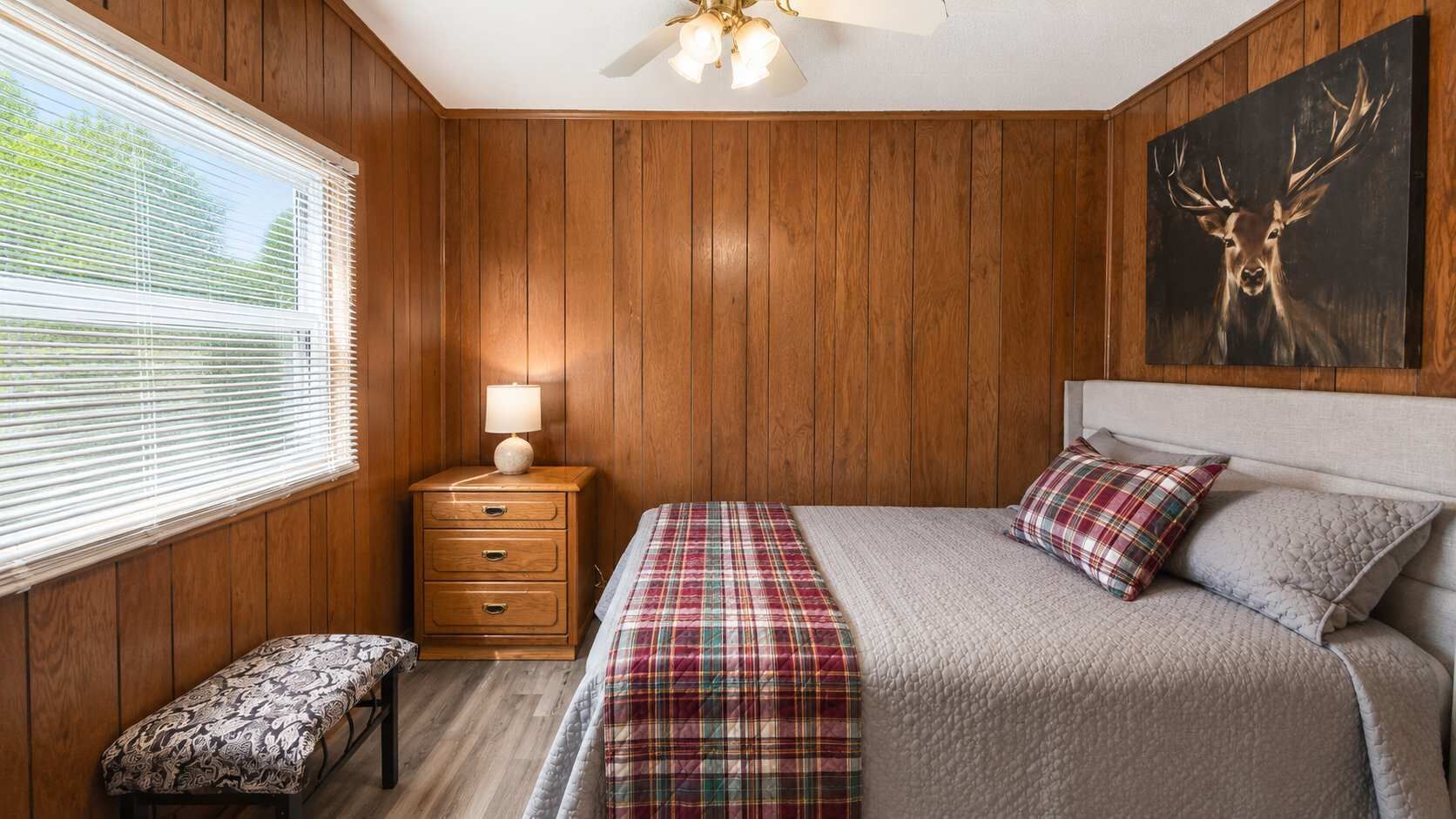 A bedroom with wooden panel walls, a bed with plaid bedding, a nightstand with a lamp, a bench, and a deer painting.