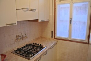 A kitchen with a gas stove, white cabinets, and a window with blinds. - Bright Flat 50m From the sea (San Michele al Tagliamento)