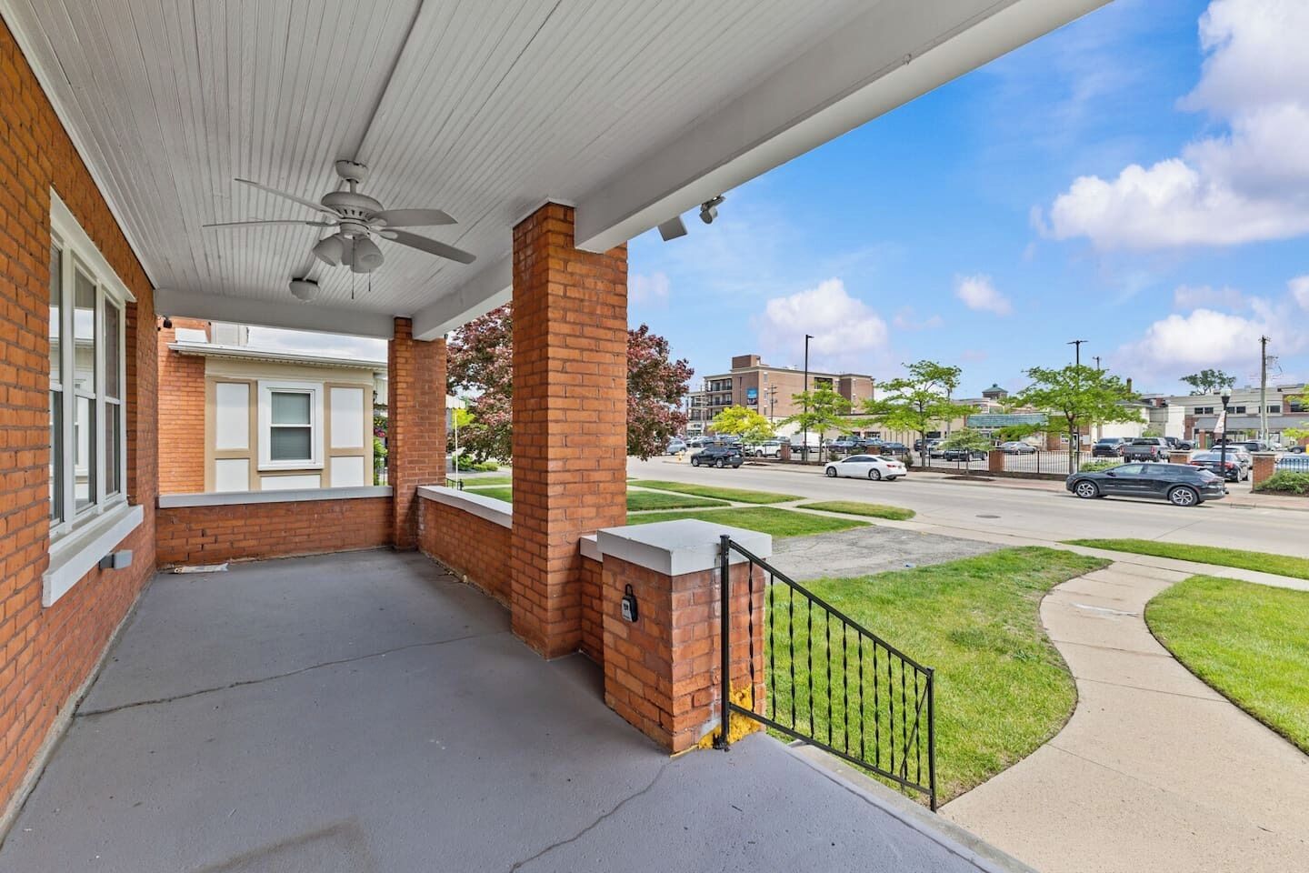 A covered porch with a ceiling fan, brick columns, and a view of a street with cars and buildings.