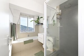 A modern bathroom with a glass-enclosed shower, a bathtub, and a window with a city view. - Serene Penthouse with Iconic Views + Private Pool (Cape Town)