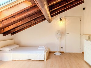 Ein Schlafzimmer mit schrägem Dach, einem Bett, einem Standventilator, einer weißen Tür und einer Kommode.