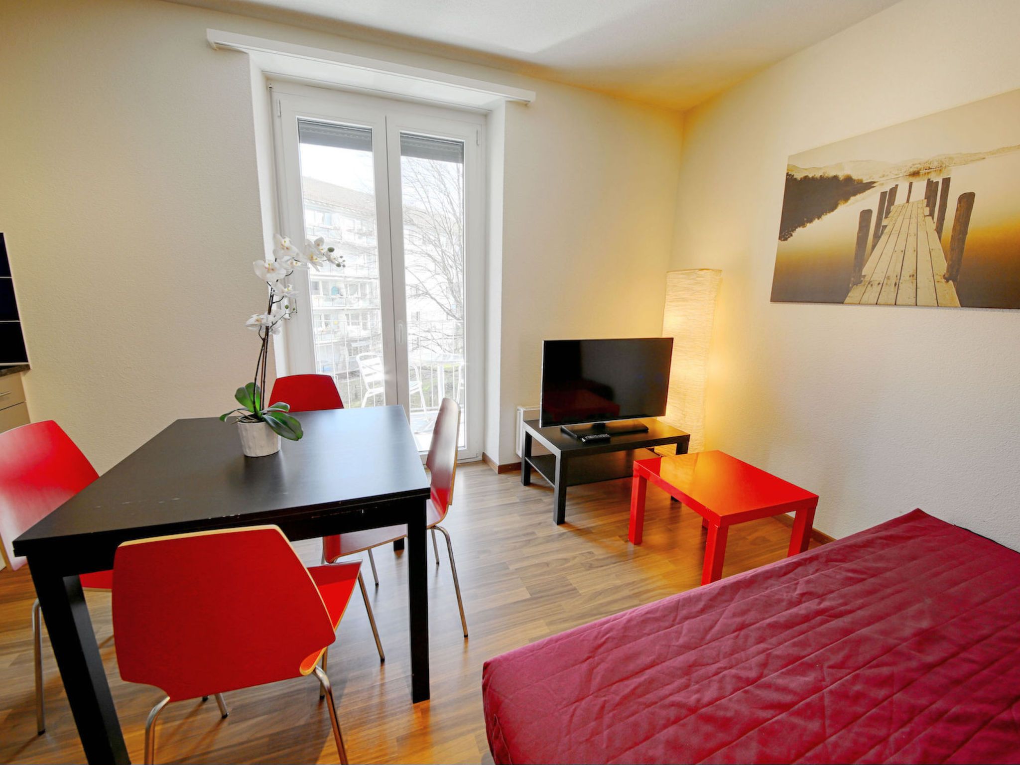 A hotel room with a bed, a TV, a desk with a chair, a small table, and a window with a view of buildings.