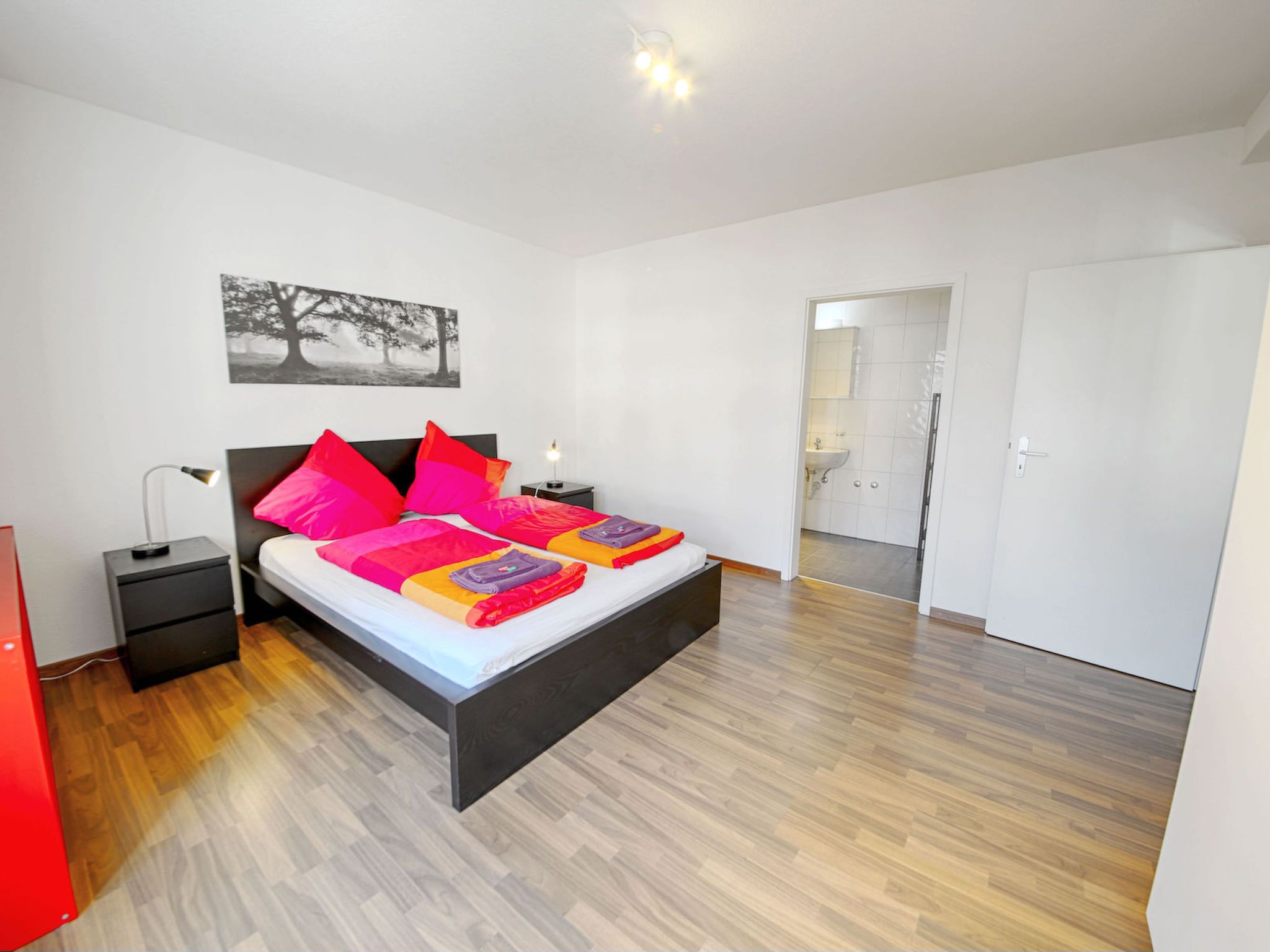 A modern bedroom with a bed, bedside tables, a nightstand, a red cabinet, and a bathroom visible through an open door.