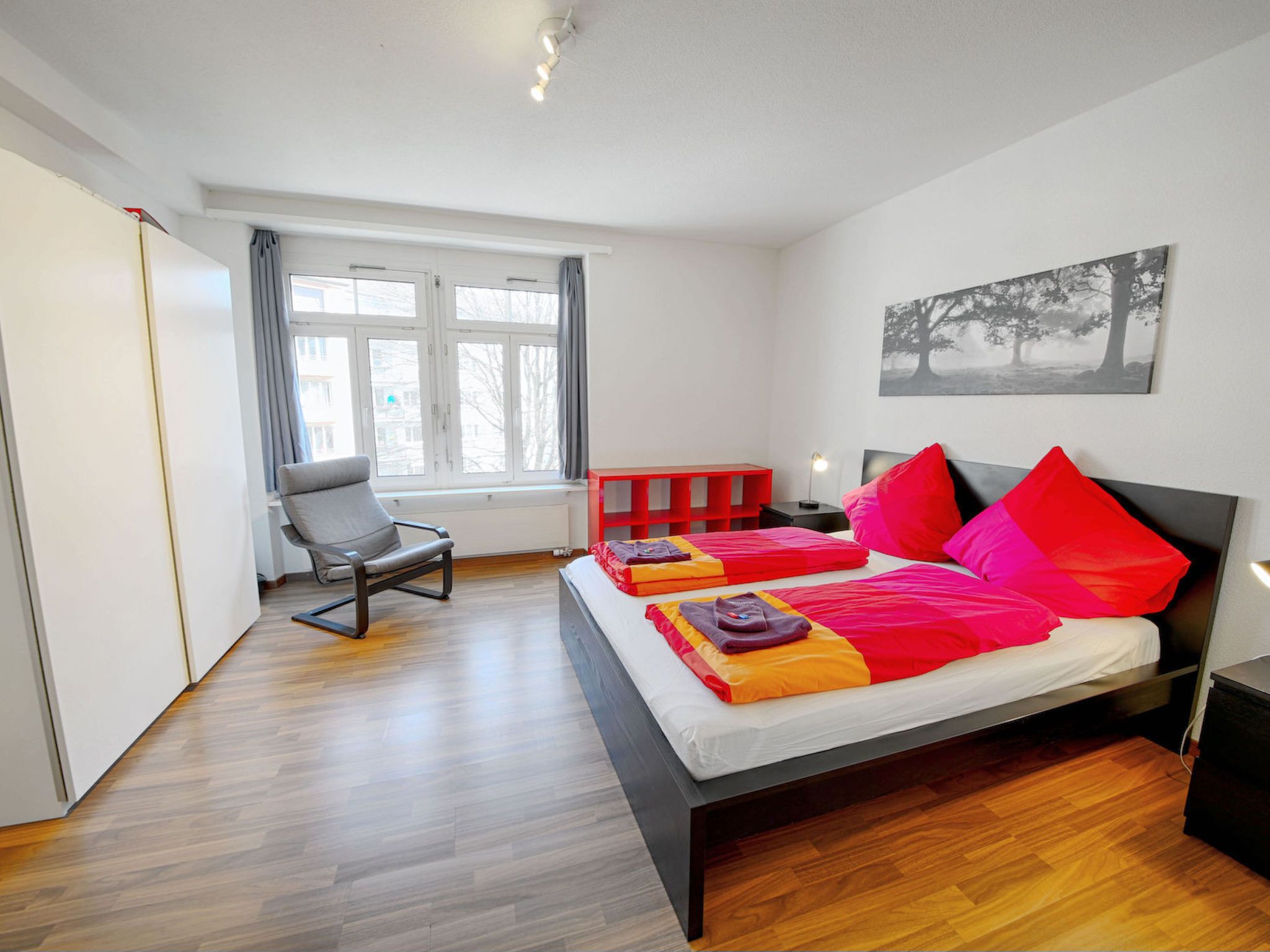 Une chambre à coucher moderne avec un grand lit, une chaise, une table de chevet et une fenêtre avec des rideaux.