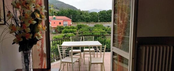 Een balkon met een tafel en stoelen, een vaas met bloemen en uitzicht op een rood gebouw en groen.