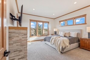 Ein Schlafzimmer mit Kamin aus Stein, einem Bett mit grau-weißer Bettwäsche, zwei Fenstern und einem hölzernen Kopfteil.