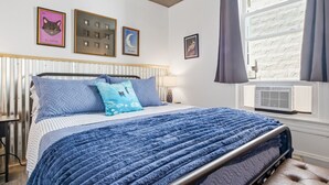 A bedroom with a bed, a nightstand, a wall-mounted air conditioner, and framed pictures on the wall. - Whimsical Haven By The Springs - Wifi | Walk to Everything (Berkeley Springs)