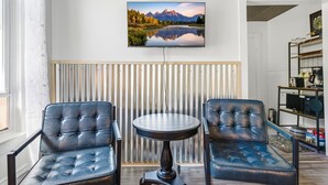 A room with two leather chairs, a round table, a mounted TV displaying a mountain landscape, a radiator, and a shelf with kitchen appliances. - Whimsical Haven By The Springs - Wifi | Walk to Everything (Berkeley Springs)