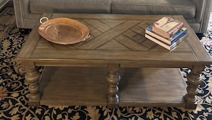 A living room with a grey sofa, patterned cushions, a wooden coffee table with books, and a patterned rug.