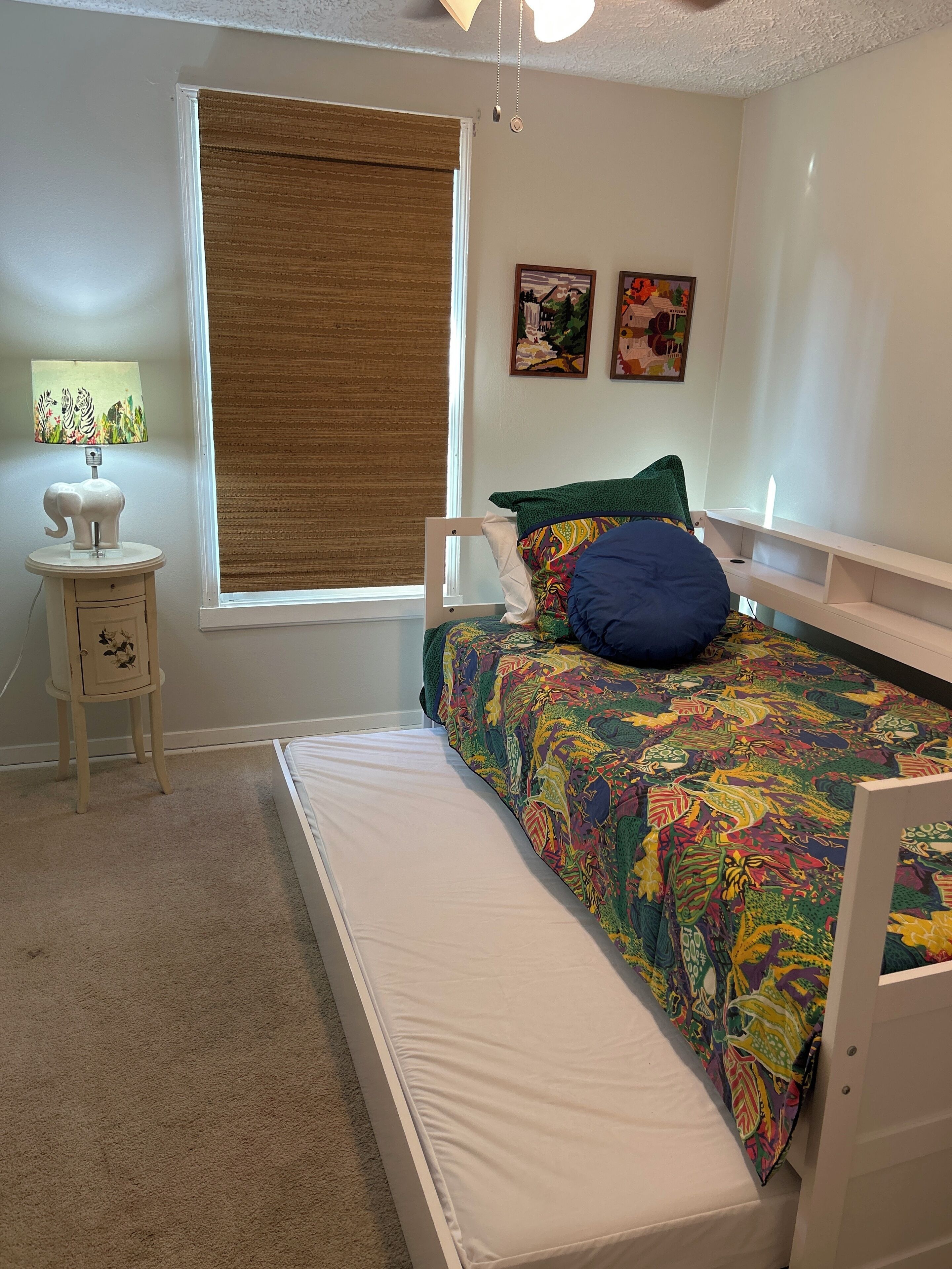 A bedroom with a bed, a nightstand, a lamp, a window with a blind, and two wall art pieces.