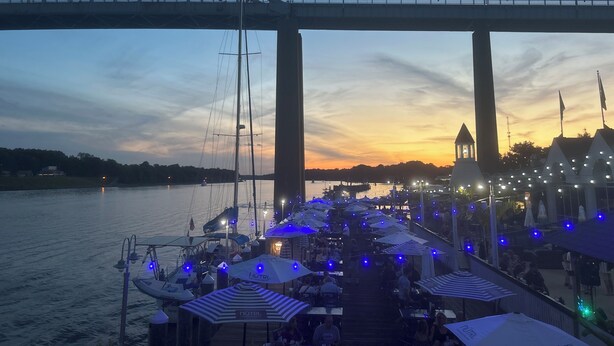 A waterfront restaurant with outdoor seating, a bridge in the background, and a sunset.
