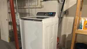A front-loading washing machine on a black platform, with a metal shelf to the right holding cleaning supplies.