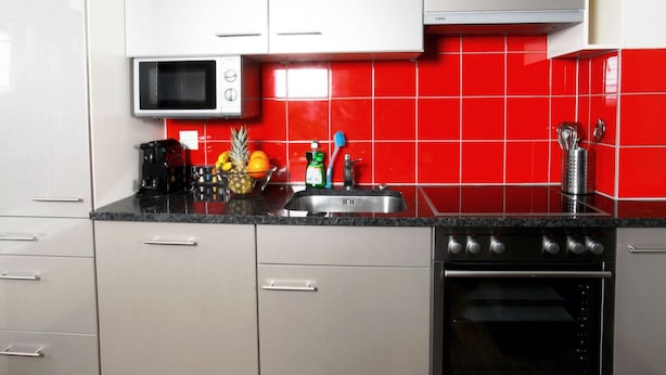 Uma cozinha com armários brancos, micro-ondas, pia e painel de azulejos vermelhos.