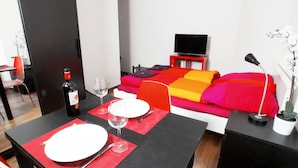 Quarto de hotel com mesa de jantar posta para dois, cama com roupa de cama colorida, televisão em um suporte e uma planta em vaso.