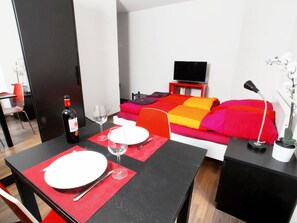 Ein Hotelzimmer mit Esstisch für zwei Personen, einem Bett mit farbenfroher Bettwäsche, einem Fernseher auf einem Beistelltisch und einer Zimmerpflanze.