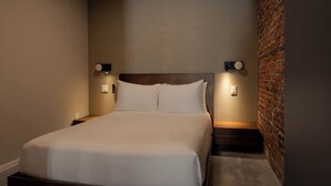 A neatly made bed with a wooden headboard, two bedside tables with wall-mounted lamps, and a brick wall on one side. - Sosuite | Deluxe Queen Room w Desk (Philadelphia)