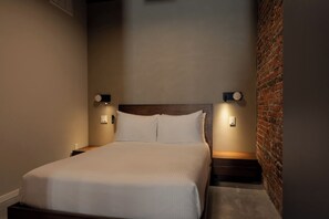 A neatly made bed with a wooden headboard, two bedside tables with wall-mounted lamps, and a brick wall on one side. - Sosuite | Deluxe Queen Room w Desk (Philadelphia)