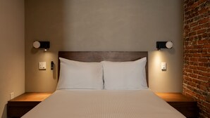 A neatly made bed with a wooden headboard, two bedside tables with wall-mounted lamps, and a brick wall on the right. - Sosuite | Deluxe Queen Room w Desk (Philadelphia)