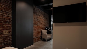 A room with a brick wall, a white chair, a flat-screen TV, and a power outlet. - Sosuite | Deluxe Queen Room w Desk (Philadelphia)