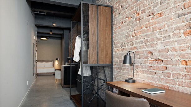 A modern room with a brick wall, a wooden desk, a metal partition, and a white sofa. - Sosuite | Deluxe Queen Room w Desk (Philadelphia)