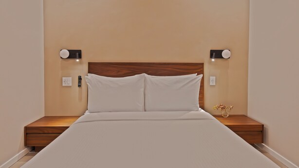 Una cama bien hecha con cabecera de madera, dos mesitas de noche con lámparas y un aplique en la pared.