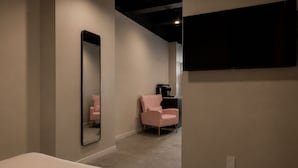 Un dormitorio con cama, espejo, una silla rosa y televisor montado en la pared.