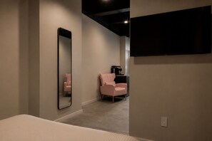 Um quarto com cama, espelho, uma cadeira rosa e televisão fixada na parede.